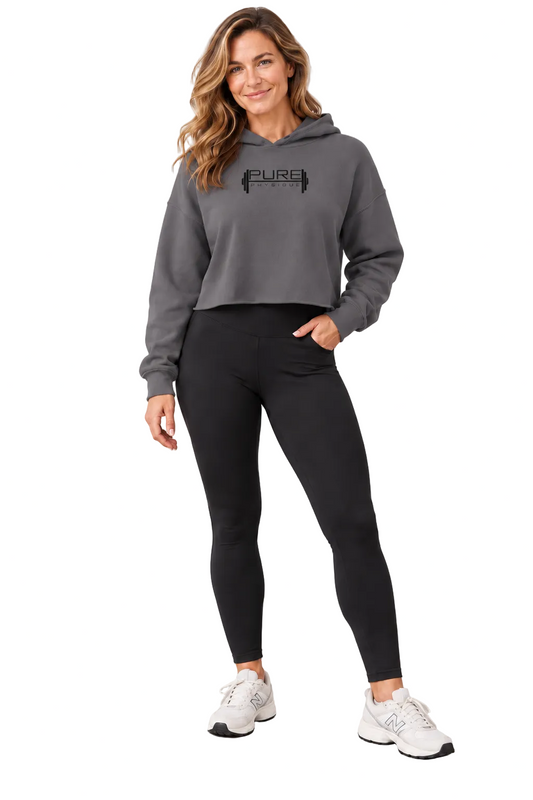 A woman modeling a dark gray cropped hoodie with a black 'PURE' graphic on the chest, paired with black leggings and white sneakers, standing in a bright studio setting.